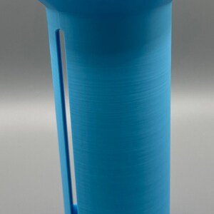 May include: A light blue plastic dispenser for storing and dispensing plastic wrap. The dispenser has a round base and a cylindrical body with a slot for the wrap.