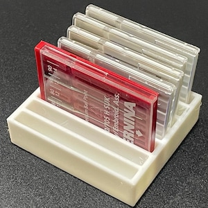 May include: A white plastic organiser with two compartments holds multiple packs of needles. The packs are labelled "BERNINA" and "0/705 H-SUK / Embroid. Ass."