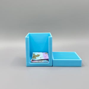 May include: A light blue plastic storage box with a separate tray. The box has a small opening on the front and the tray has a series of slots. A stack of colorful patterned paper is inside the box.