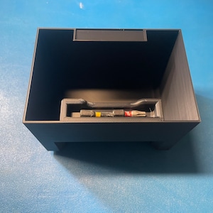 Puede incluir: Una caja de almacenamiento de plástico negro con un compartimento para guardar herramientas pequeñas. La caja tiene una tapa y una pequeña manija en la parte superior. La caja está vacía.