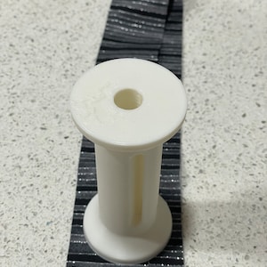 Op de afbeelding: Witte plastic spoel met een gat in het midden. De spoel ligt op een zwart-zilveren gestreept lint.