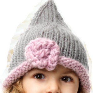 Könnte beinhalten: Graue Strickmütze mit spitzer Spitze und rosa Blumenakzent. Der Hut hat einen rosa Rand und wird von einem jungen Kind mit lockigem blondem Haar getragen. Ein gemütliches Accessoire.