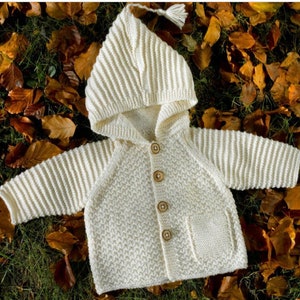 May include: Cream-colored knitted baby cardigan with a hood and wooden buttons. The hooded sweater has a textured stitch pattern on the body and ribbed knit sleeves. A small pocket is on the lower right side. The hood has a tassel.