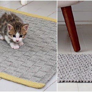 May include: A gray rectangular knitted rug with a yellow border. A small kitten with brown and white markings is on the rug. The rug has a textured, woven pattern. The rug is approximately 24 inches long.