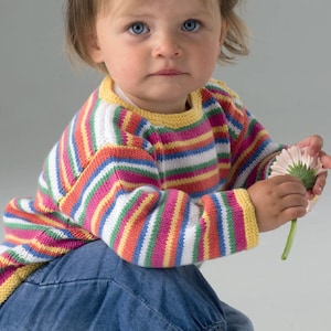 Könnte beinhalten: Ein Kind trägt einen bunten gestreiften Pullover mit gelbem Kragen und Bündchen, kombiniert mit einer blauen Jeans. Der Pullover hat horizontale Streifen in verschiedenen Farben. Das Kind hält eine rosa Blume mit grünem Stiel.