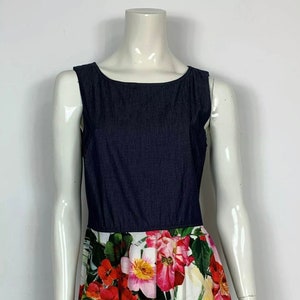 Può includere: Un abito senza maniche con un corpetto in denim blu scuro e una gonna a fiori bianca. La gonna presenta una varietà di fiori rosa, rossi e verdi.