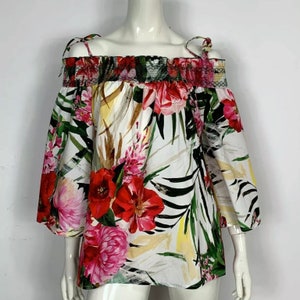 Può includere: Una blusa bianca a spalle scoperte con stampa floreale che presenta fiori e foglie rosa, rossi e verdi. La blusa ha un collo a volant e una vestibilità ampia.