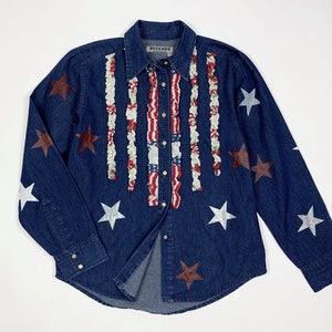 K&ouml;nnte beinhalten: Ein blaues Jeanshemd mit wei&szlig;em und rotem R&uuml;schenbesatz an der Vorderseite. Das Hemd hat silberne und rote Stern-Akzente.