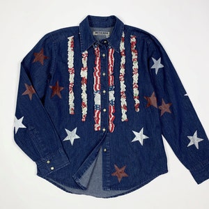 K&ouml;nnte beinhalten: Ein blaues Jeanshemd mit roten, wei&szlig;en und blauen R&uuml;schenbesatz und silbernen und roten Stern-Applikationen. Das Hemd hat einen Knopfleisten vorne und lange &Auml;rmel. Das Etikett auf dem Hemd lautet "Beckard".