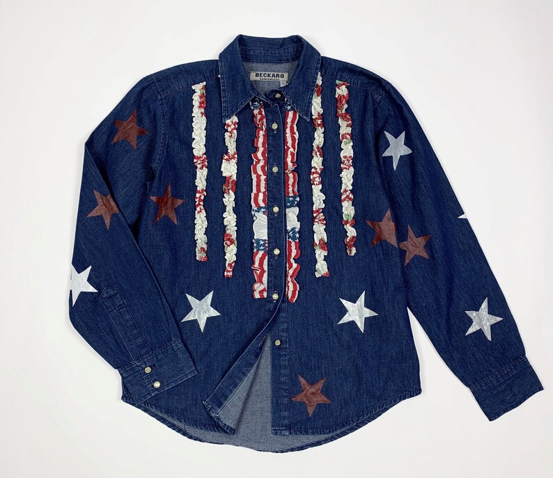 K&ouml;nnte beinhalten: Ein blaues Jeanshemd mit roten, wei&szlig;en und blauen R&uuml;schenbesatz und silbernen und roten Stern-Applikationen. Das Hemd hat einen Knopfleisten vorne und lange &Auml;rmel. Das Etikett auf dem Hemd lautet "Beckard".