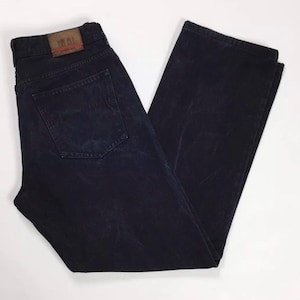 Pode incluir: Jeans de ganga azul marinho escuro com um design clássico de cinco bolsos. Os jeans têm um corte de perna reto e um remendo de couro castanho na cintura traseira. Os jeans estão dobrados e colocados sobre uma superfície branca.