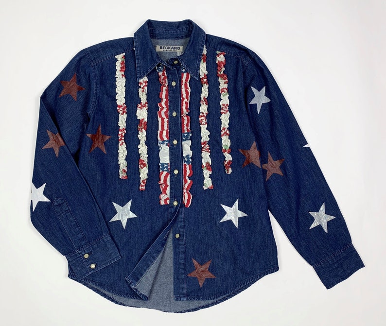 K&ouml;nnte beinhalten: Ein blaues Jeanshemd mit einem roten, wei&szlig;en und blauen R&uuml;schenmuster und silbernen und braunen Sternen. Das Hemd hat einen Knopfleisten vorne und lange &Auml;rmel. Das Etikett auf dem Hemd lautet "Beckard Collection".