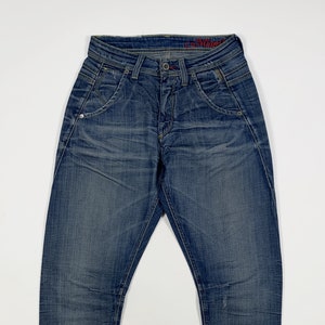 Può includere: Un paio di jeans blu denim con un lavaggio sbiadito e un aspetto leggermente rovinato. I jeans hanno una gamba dritta e una vita media. Il nome del marchio "Mavi" è ricamato sulla cintura posteriore.