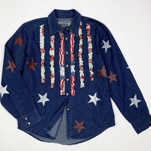 K&ouml;nnte beinhalten: Ein blaues Jeanshemd mit einem roten, wei&szlig;en und blauen R&uuml;schenmuster und silbernen und braunen Sternen. Das Hemd hat einen Knopfleisten vorne und lange &Auml;rmel. Das Etikett auf dem Hemd lautet "Beckard Collection".