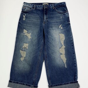 Può includere: Culotte in denim blu con dettagli strappati. I pantaloni a gamba larga presentano strappi e lacerazioni sulla parte anteriore delle gambe. I risvolti sono ripiegati. I jeans hanno un classico design a cinque tasche.