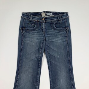 Può includere: Jeans blu in denim con chiusura a bottone e cuciture decorative intorno alle tasche. I jeans hanno un lavaggio sbiadito e un leggero svasatura sul fondo. I jeans hanno una vita media e un classico design a cinque tasche.