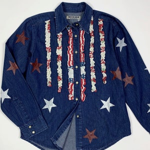 K&ouml;nnte beinhalten: Ein blaues Jeanshemd mit rotem, wei&szlig;em und blauem R&uuml;schenbesatz an der Vorderseite. Das Hemd hat silberne und braune Sterne auf der Vorderseite und den &Auml;rmeln. Das Hemd hat ein Etikett mit der Aufschrift "Beckard" auf der Vorderseite.