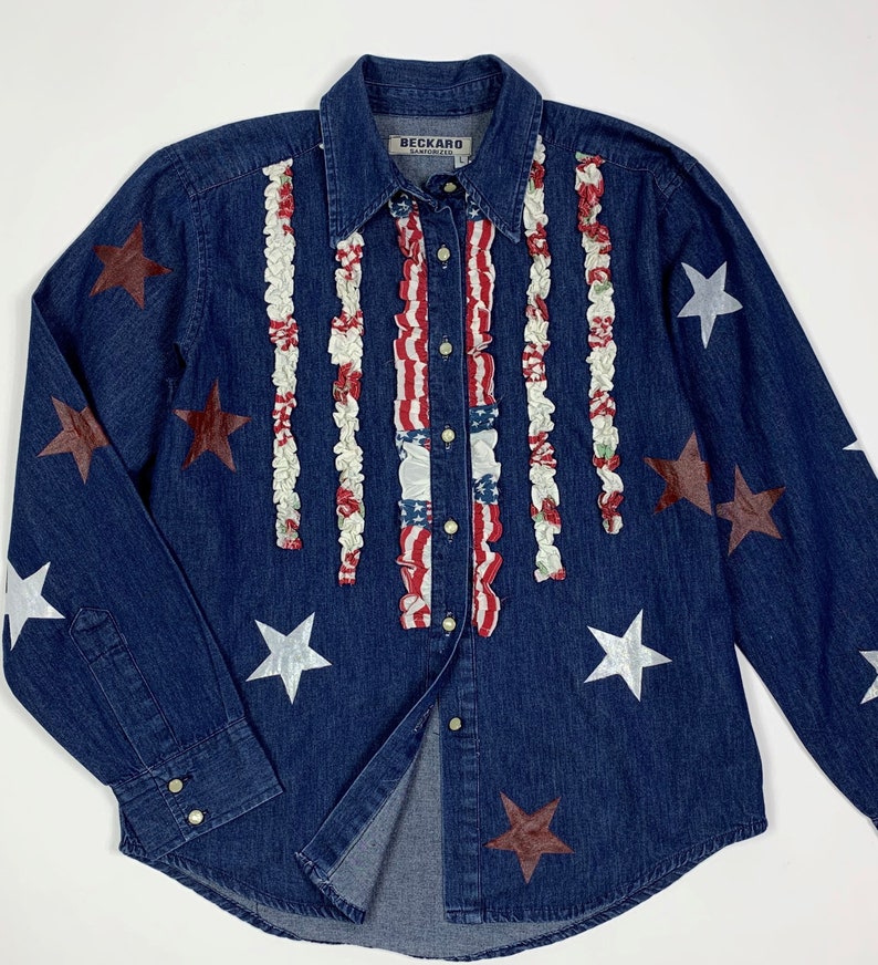 K&ouml;nnte beinhalten: Ein blaues Jeanshemd mit rotem, wei&szlig;em und blauem R&uuml;schenbesatz an der Vorderseite. Das Hemd hat silberne und braune Sterne auf der Vorderseite und den &Auml;rmeln. Das Hemd hat ein Etikett mit der Aufschrift "Beckard" auf der Vorderseite.