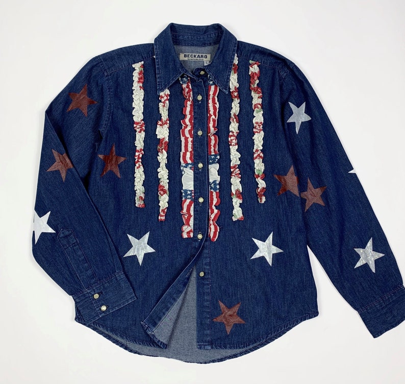 K&ouml;nnte beinhalten: Ein blaues Jeanshemd mit wei&szlig;em und rotem R&uuml;schenbesatz an der Vorderseite. Das Hemd hat silberne und rote Stern-Akzente.