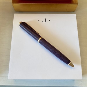 May include: A white notepad with the letter "J" written on it. A purple pen with a gold trim is lying on the notepad.