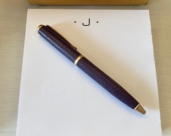 May include: A white notepad with the letter "J" written on it. A purple pen with a gold trim is lying on the notepad.