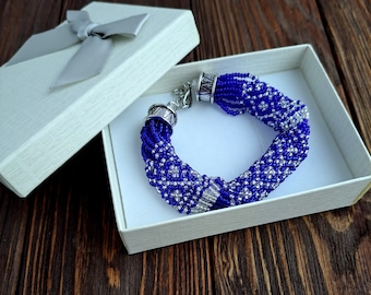Può includere: Un braccialetto di perline blu e argento in una scatola regalo bianca. Il braccialetto è fatto di piccole perline e ha una chiusura argentata.