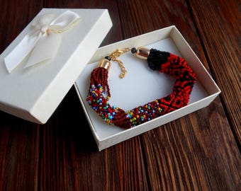 Può includere: Un braccialetto di perline in una confezione regalo bianca con un fiocco color crema. Il braccialetto presenta perline rosse, nere e multicolori. La scatola è aperta, rivelando il gioiello all'interno. Il braccialetto è fatto di piccole perline colorate.