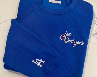 May include: A royal blue sweatshirt with white embroidered lettering that says "Los Dodgers" and a baseball graphic. The left sleeve has a white embroidered "LA" logo.