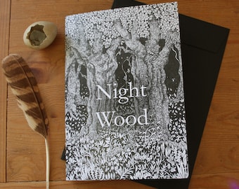 May include: A card with the words "Night Wood" and a black and white forest illustration. A black envelope is behind the card. A brown and white feather and a small, light-coloured stone are on a wooden surface.