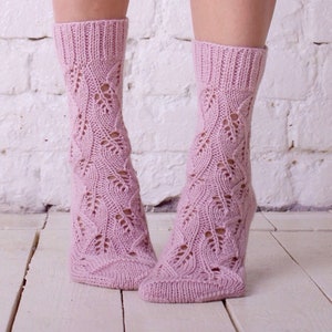 May include: A pair of pink knitted socks with a lacy pattern. The socks are shown on a pair of legs against a white brick wall background.