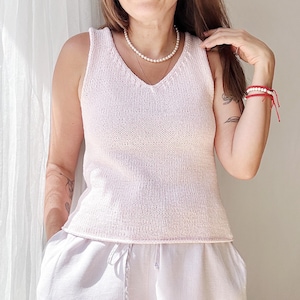 以下が含まれることがあります： Vネックとパールネックレスが付いた薄いピンクのノースリーブニットトップ。トップは白いリネンショーツと組み合わせています。