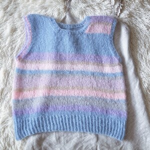 Puede incluir: Un chaleco de punto en colores pastel con rayas horizontales azules, rosas, moradas y grises. La parte superior sin mangas tiene un cuello redondo y está hecha de un hilo suave y texturizado. El chaleco está sobre un camisola gris claro.