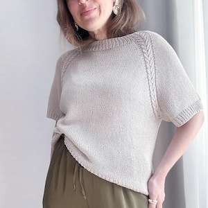 Può includere: Un top di maglia beige chiaro con un dettaglio a treccia sulle maniche. Il top ha una scollatura rotonda e maniche corte. La modella indossa il top con pantaloni verde oliva.