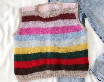 Puede incluir: Un chaleco de punto a rayas multicolores en tonos rosa, azul, amarillo, verde y marrón. El chaleco es sin mangas y tiene cuello redondo. Unos vaqueros de mezclilla azul claro están al lado del chaleco. Un broche y anillos están en el bolsillo de los vaqueros.