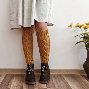 May include: A pair of mustard yellow knitted knee-high socks with a cable knit pattern. The socks are worn with brown leather ankle boots.
