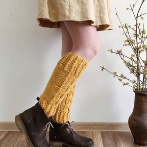 May include: A pair of yellow knitted leg warmers with a cable knit pattern. The leg warmers are worn over brown leather boots.
