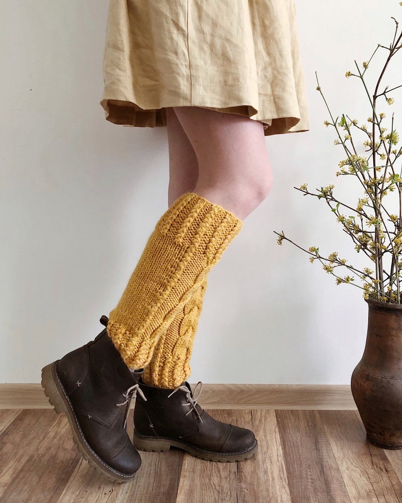 May include: A pair of yellow knitted leg warmers with a cable knit pattern. The leg warmers are worn over brown leather boots.