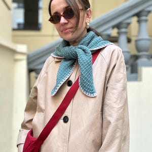 Op de afbeelding: Een persoon draagt een beige trenchcoat met zwarte knopen, een turquoise gebreide sjaal met witte rand en een rode crossbodytas. De persoon draagt ook een zonnebril en oorringen.