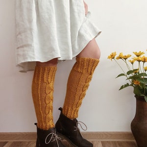 May include: A pair of mustard yellow knitted knee-high socks with a cable knit pattern. The socks are worn with brown leather boots and a white linen skirt.