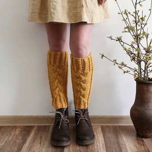 May include: A pair of mustard yellow knitted leg warmers with a cable knit pattern. The leg warmers are worn over brown ankle boots.