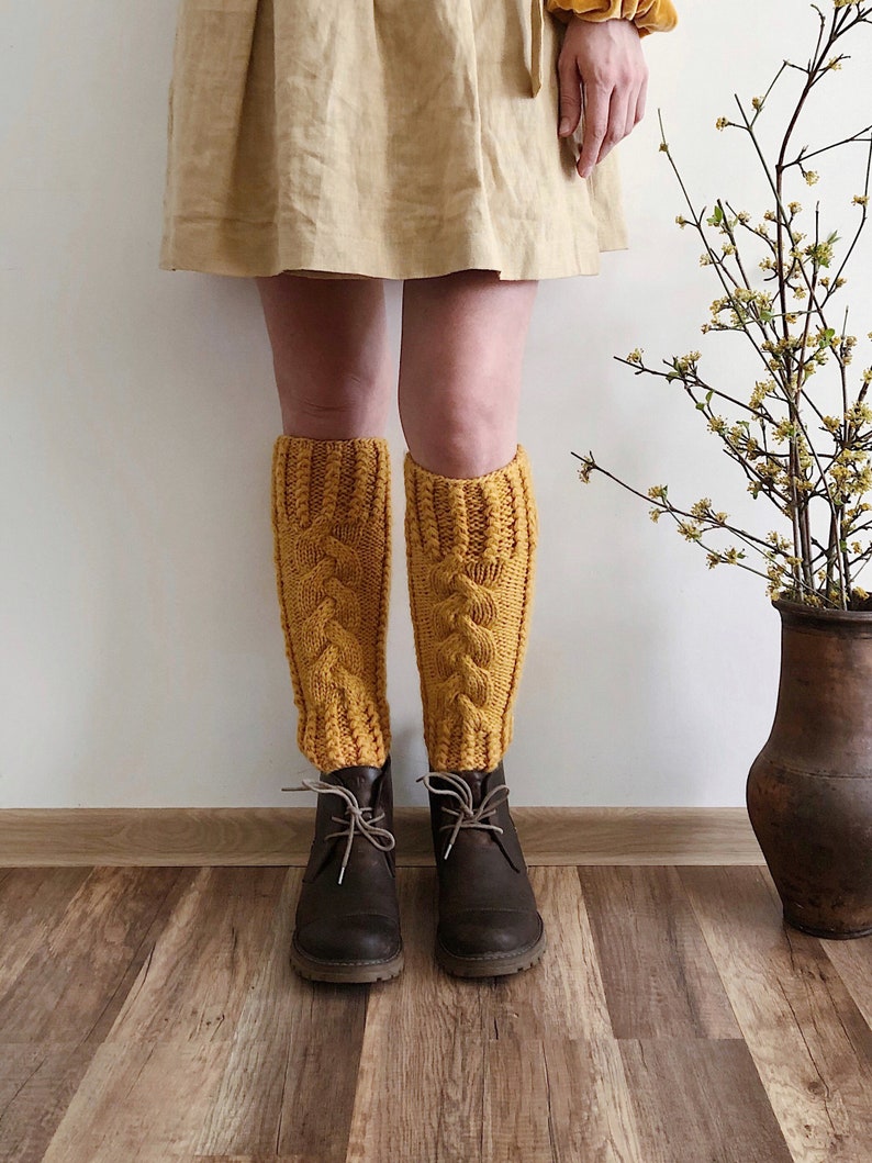 May include: A pair of mustard yellow knitted leg warmers with a cable knit pattern. The leg warmers are worn over brown ankle boots.