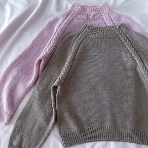 Puede incluir: Dos suéteres de punto, uno lila claro y otro gris, se muestran sobre una superficie blanca. Ambos suéteres tienen mangas largas, puños acanalados y un cuello acanalado. Los hombros presentan un diseño trenzado decorativo.