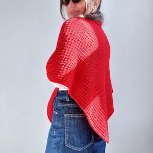 Puede incluir: Un chal rojo brillante tejido con un patrón texturizado. El chal está drapeado sobre los hombros, creando un look elegante y cálido. El modelo lleva vaqueros azules y gafas de sol.