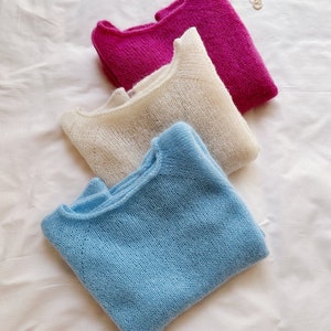 Può includere: Tre maglioni di lana in diversi colori: un maglione blu chiaro, un maglione color crema e un maglione rosa acceso. I maglioni sono piegati e impilati uno sopra l'altro su una superficie bianca.