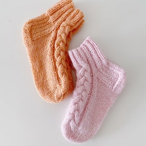 Può includere: Due paia di calzini a maglia, uno di colore arancio chiaro e l'altro di colore rosa chiaro. Entrambi i calzini hanno un motivo a treccia.