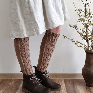 May include: A pair of brown knitted leg warmers with a cable knit pattern. The leg warmers are worn with brown leather boots.