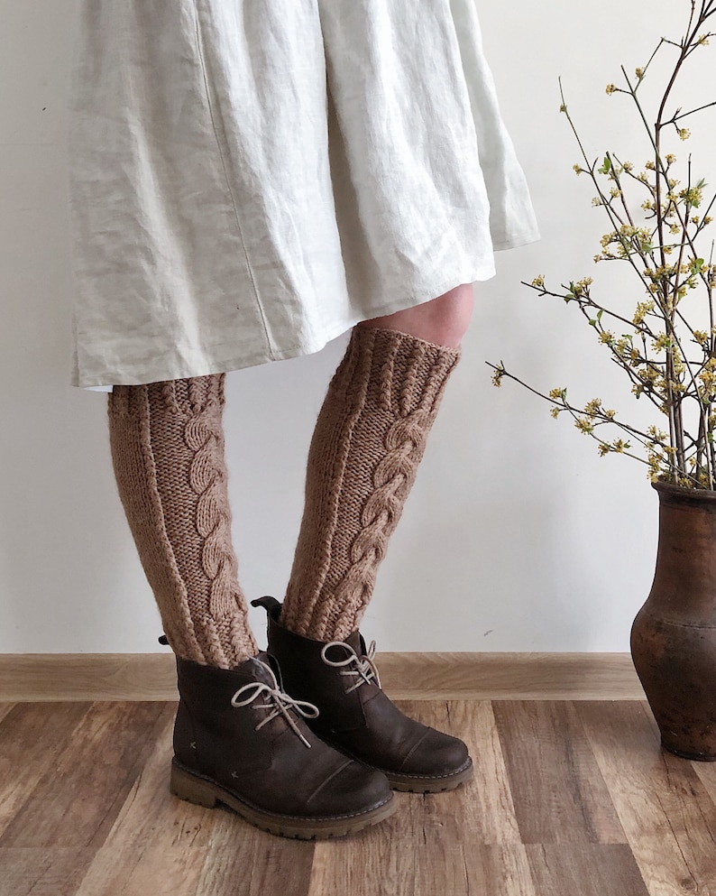 May include: A pair of brown knitted leg warmers with a cable knit pattern. The leg warmers are worn with brown leather boots.