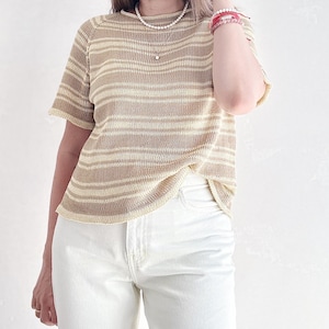 Könnte beinhalten: Eine Frau trägt ein gestreiftes, beige-weißes Strickoberteil mit kurzen Ärmeln und weiße Jeans. Sie trägt eine Perlenkette und mehrere Armbänder am Handgelenk.