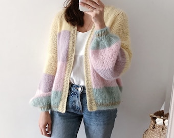 May include: A pastel-colored, open-front cardigan with a striped design. The sweater features a combination of yellow, lavender, pink, and blue horizontal stripes. The sleeves are long and voluminous. The person is wearing blue jeans and a white top.
