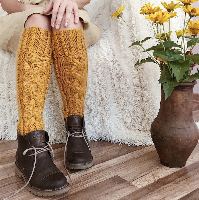 May include: A pair of mustard yellow cable knit knee-high socks with brown leather boots. The socks have a thick, chunky knit and are worn with a casual outfit.